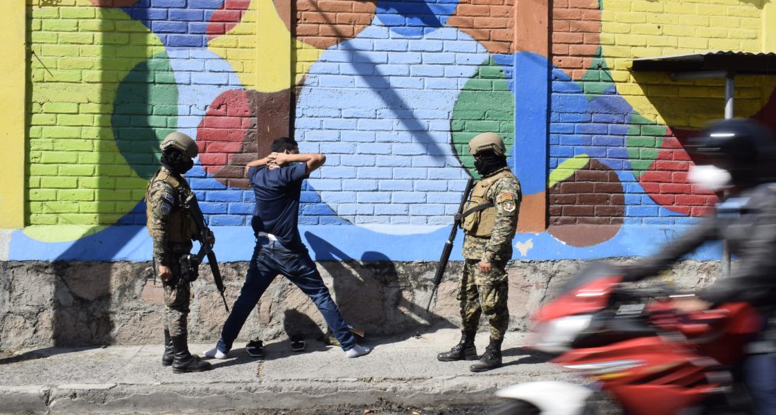 Ein Mann mit erhobenen Händen hinter dem Kopf und zwei uniformierten Militärs daneben
