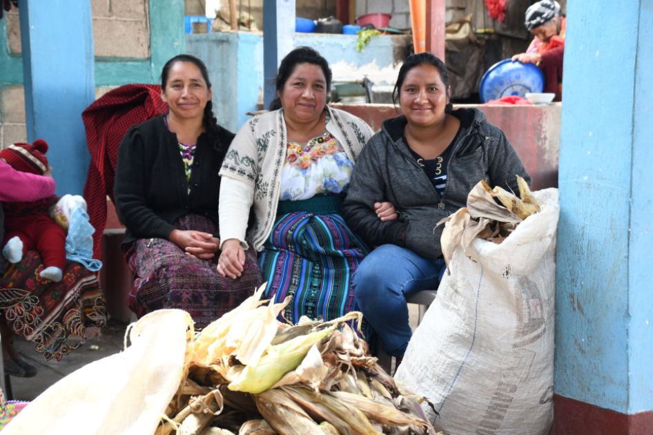Drei Teilnehmer*innen des INKOTA-Projekts Zukunft säen blicken in die Kamera. Im Vordergrund ist ihre Maisernte zu sehen.