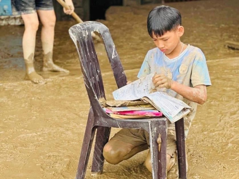 Ein Plastikstuhl steht im Wasser, davor hockt ein Junge und sortiert seine Schulsachen, die auf dem Stuhl liegen