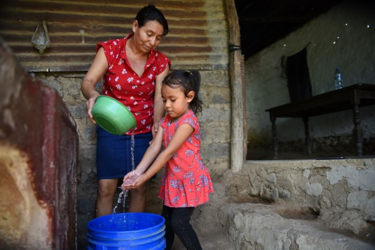 Eine Frau gießt aus einer Schüssel Wasser, daneben steht ein Kind und hält die Hände unter den Wasserstrahl, darunter eine blauer Tonne